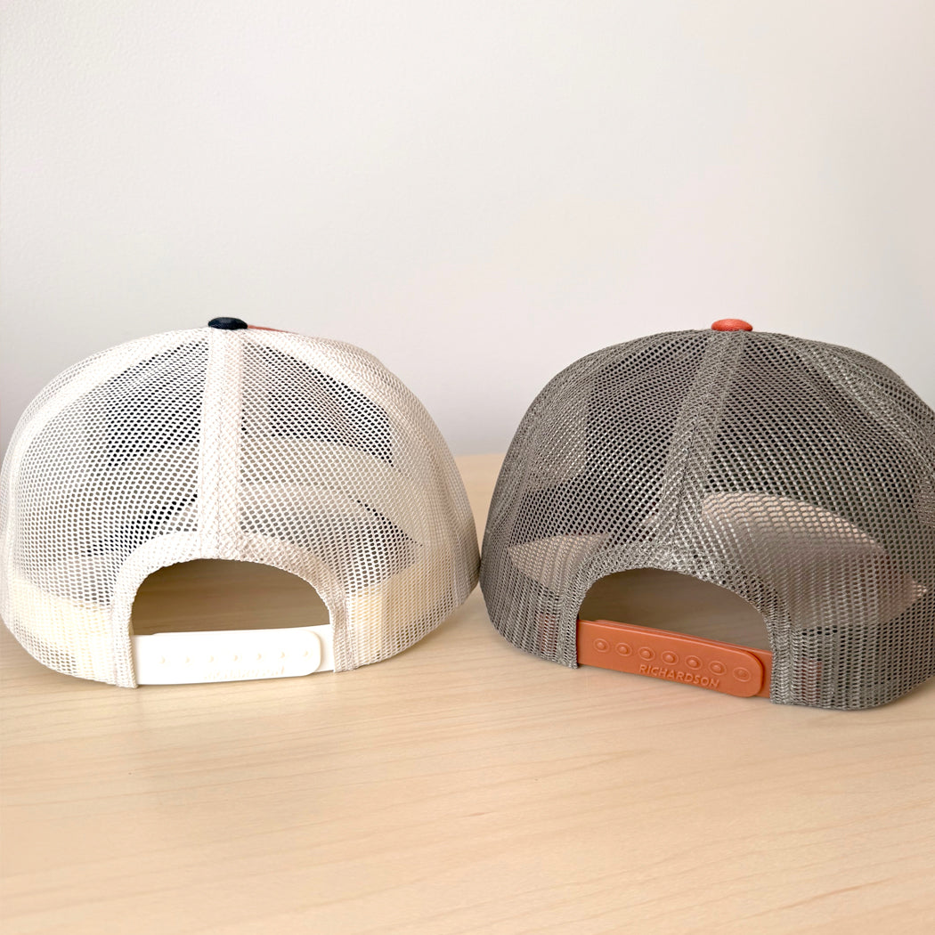 Two mesh caps, one white and one gray with orange brim, on a light wooden surface.