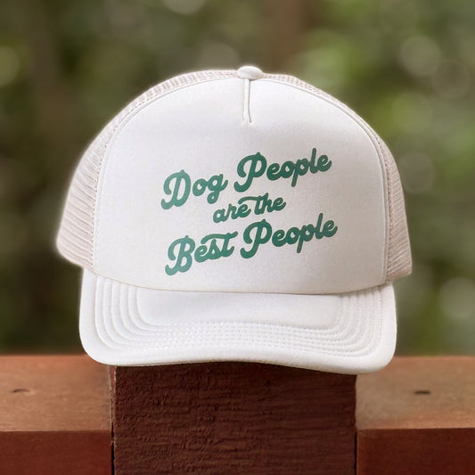 White cap with 'Dog People are the Best People' text on a wooden surface with a blurred green background