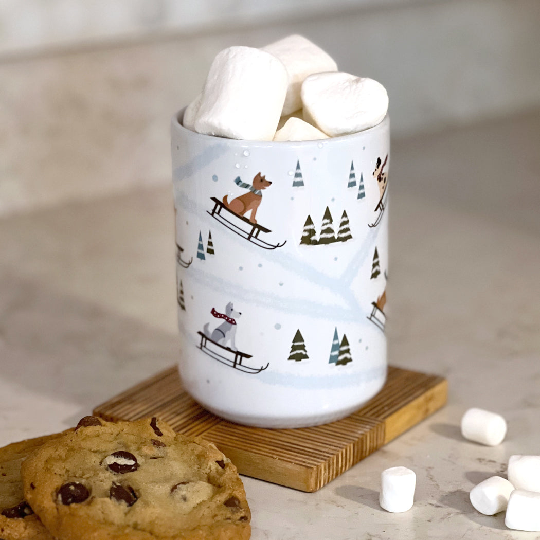 White mug with illustrated dogs on sleds, in a winter scene with pine trees and snow flakes.