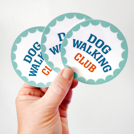 Hand holding a round vinyl Dog Walking Club sticker with teal scalloped border and bold text.