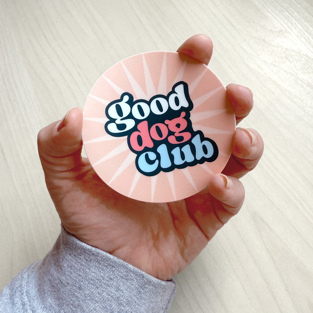 Hand holding a pink round Good Dog Club vinyl sticker with retro lettering and sunburst design.