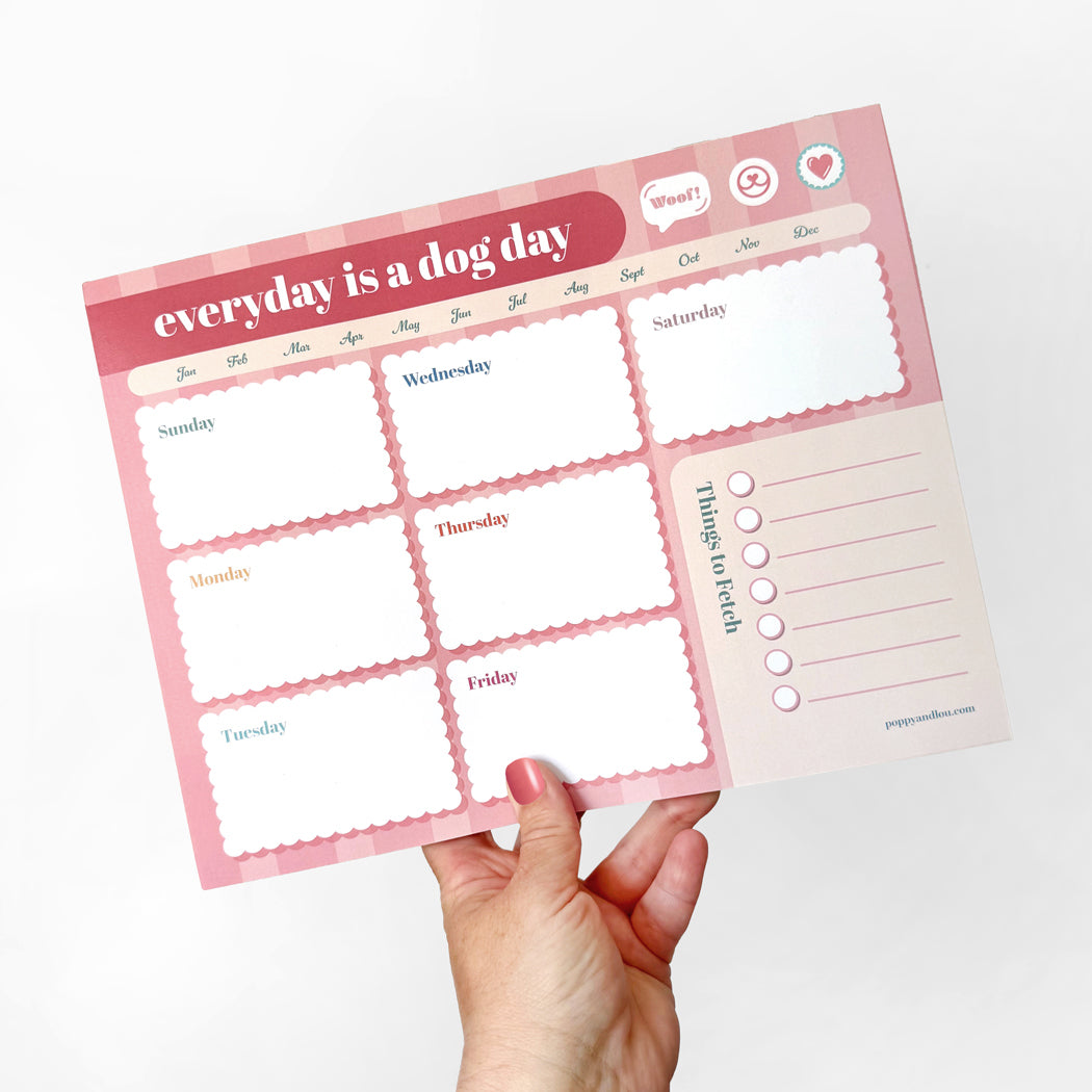 Hand holding a pink Dog Day Weekly Planner with daily boxes and to-do list, cute undated desk pad for dog lovers and organizers.
