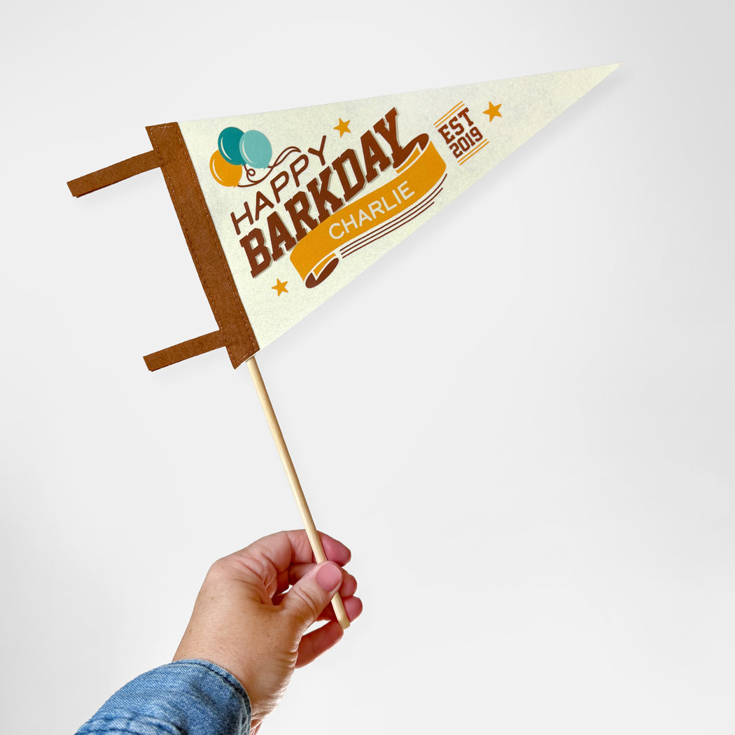 Hand holding a small flag with 'Happy Barkday Charlie' text on a white background