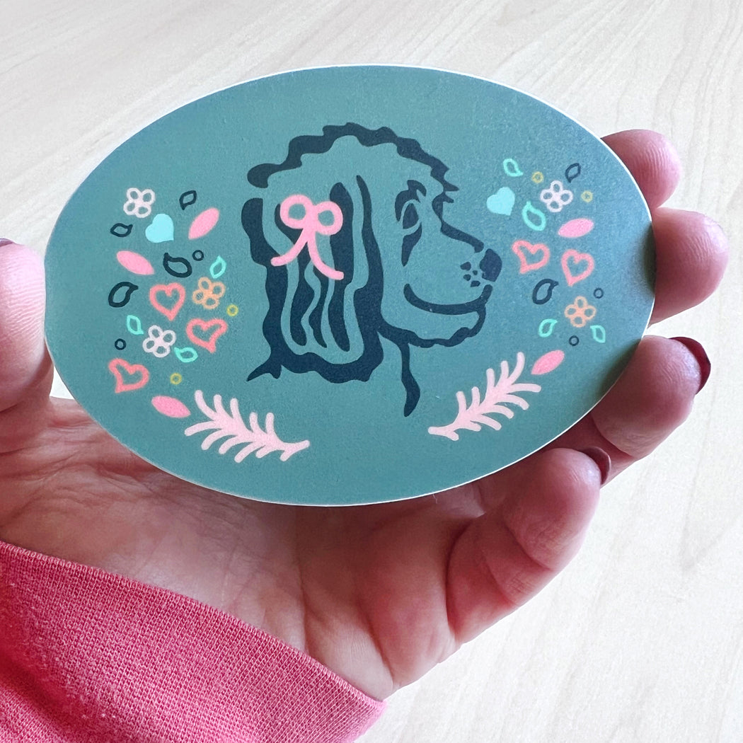 Oval sticker featuring a doodle dog with pink and blue hearts, flowers, and leaves.