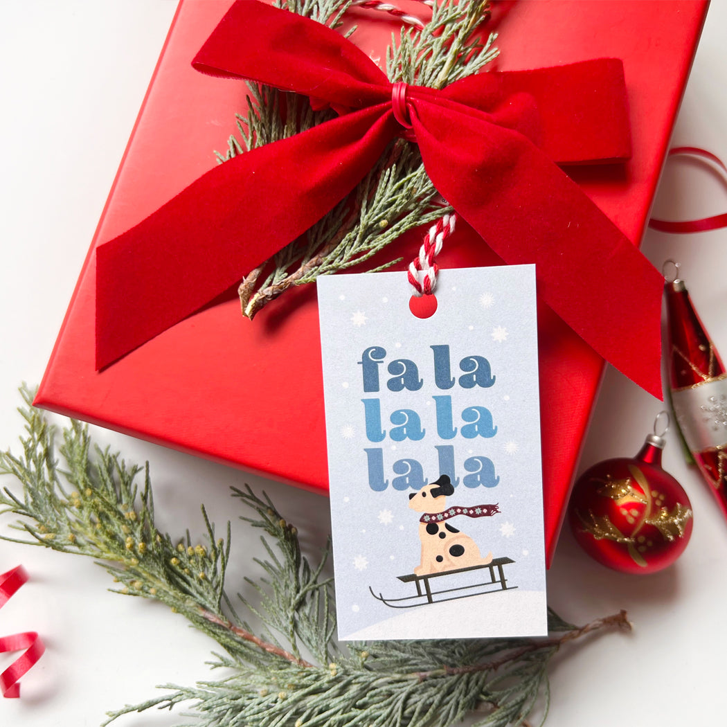 Red gift box with a festive tag featuring a dog on a sleigh and 'fa la la la la' text, surrounded by Christmas decorations.
