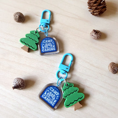 Two matching camp-themed keychains with blue carabiners, clear acrylic tags reading "CAMP CANINE," and green tree charms, arranged on a light wood surface with pinecones around them.