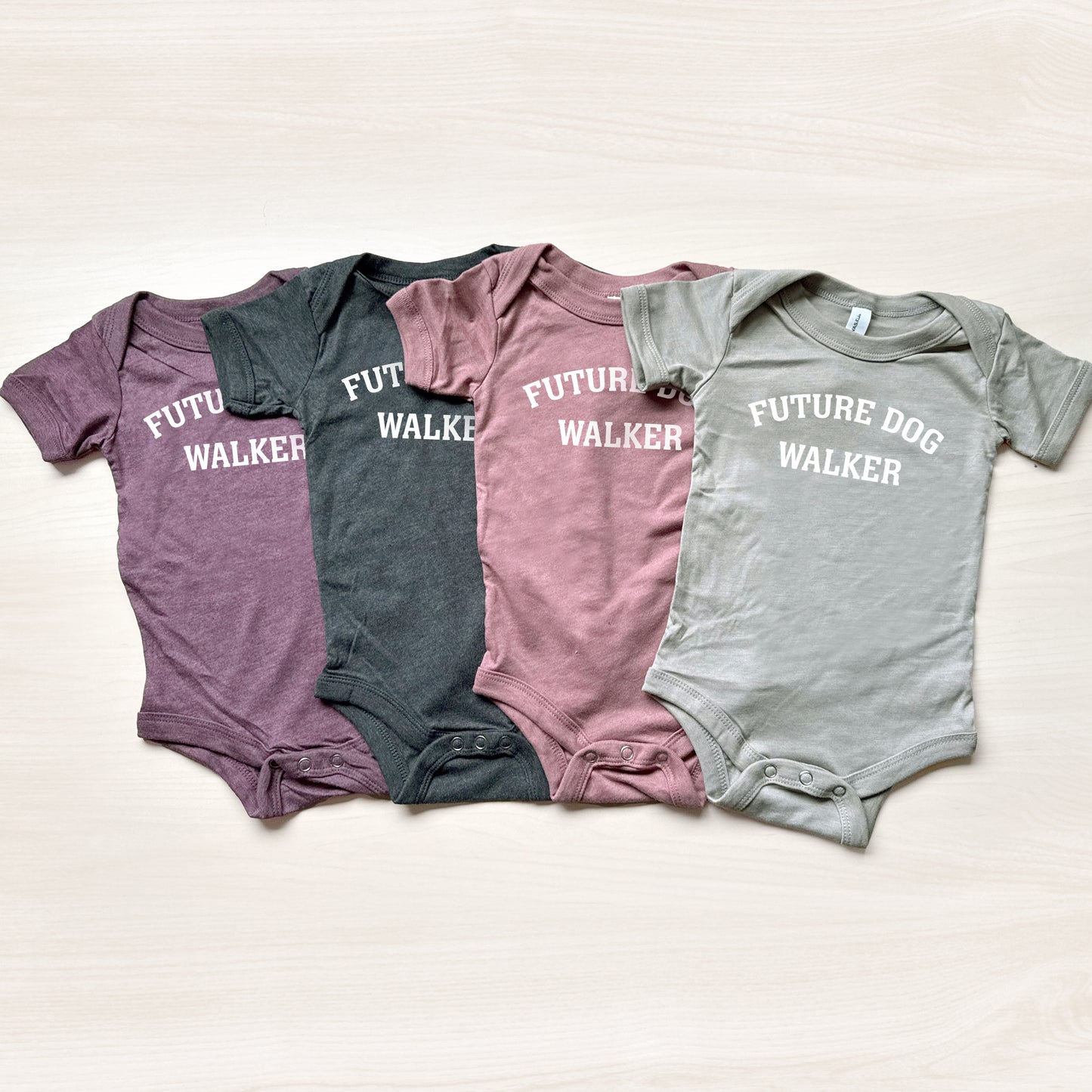 Four baby onesies in different colors with 'FUTURE DOG WALKER' text on a light background