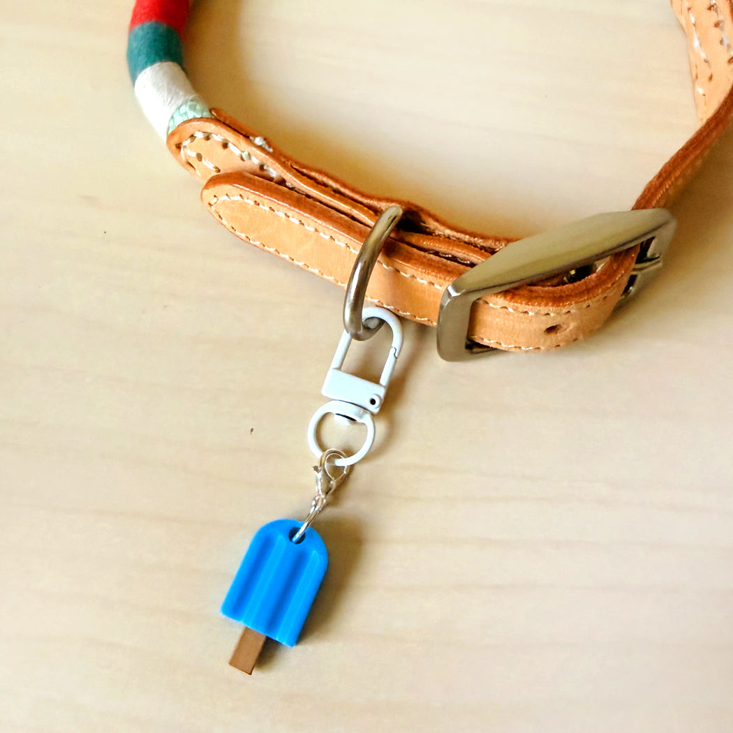A close-up of a tan leather collar with a silver metal buckle and a small blue popsicle-shaped charm hanging from a silver clasp, placed on a light wood surface.