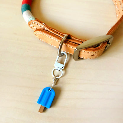 A close-up of a tan leather collar with a silver metal buckle and a small blue popsicle-shaped charm hanging from a silver clasp, placed on a light wood surface.