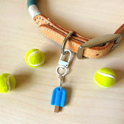 A close-up of a tan leather dog collar with a silver metal clasp and a small blue popsicle-shaped USB flash drive charm, styled on a light wood surface with three yellow tennis balls.