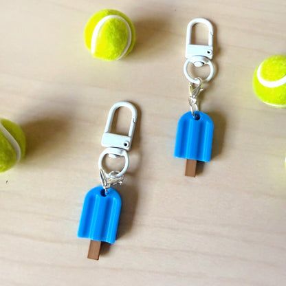 Two blue popsicle-shaped keychains with white lobster clamps, displayed on a light wood surface with tennis balls as props.