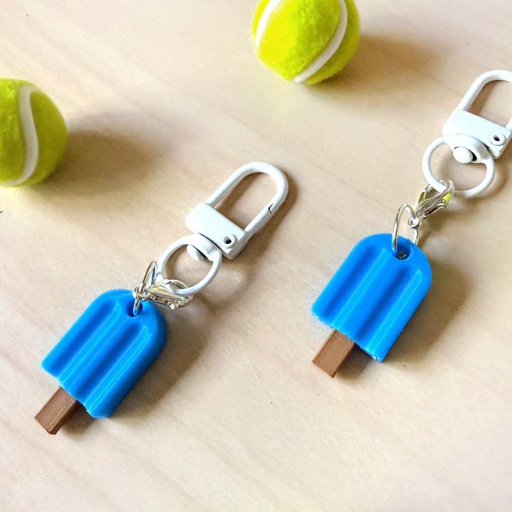 Two popsicle-shaped keychains in bright blue with brown wooden sticks and white carabiners, displayed on a light wood surface with tennis balls in the background.