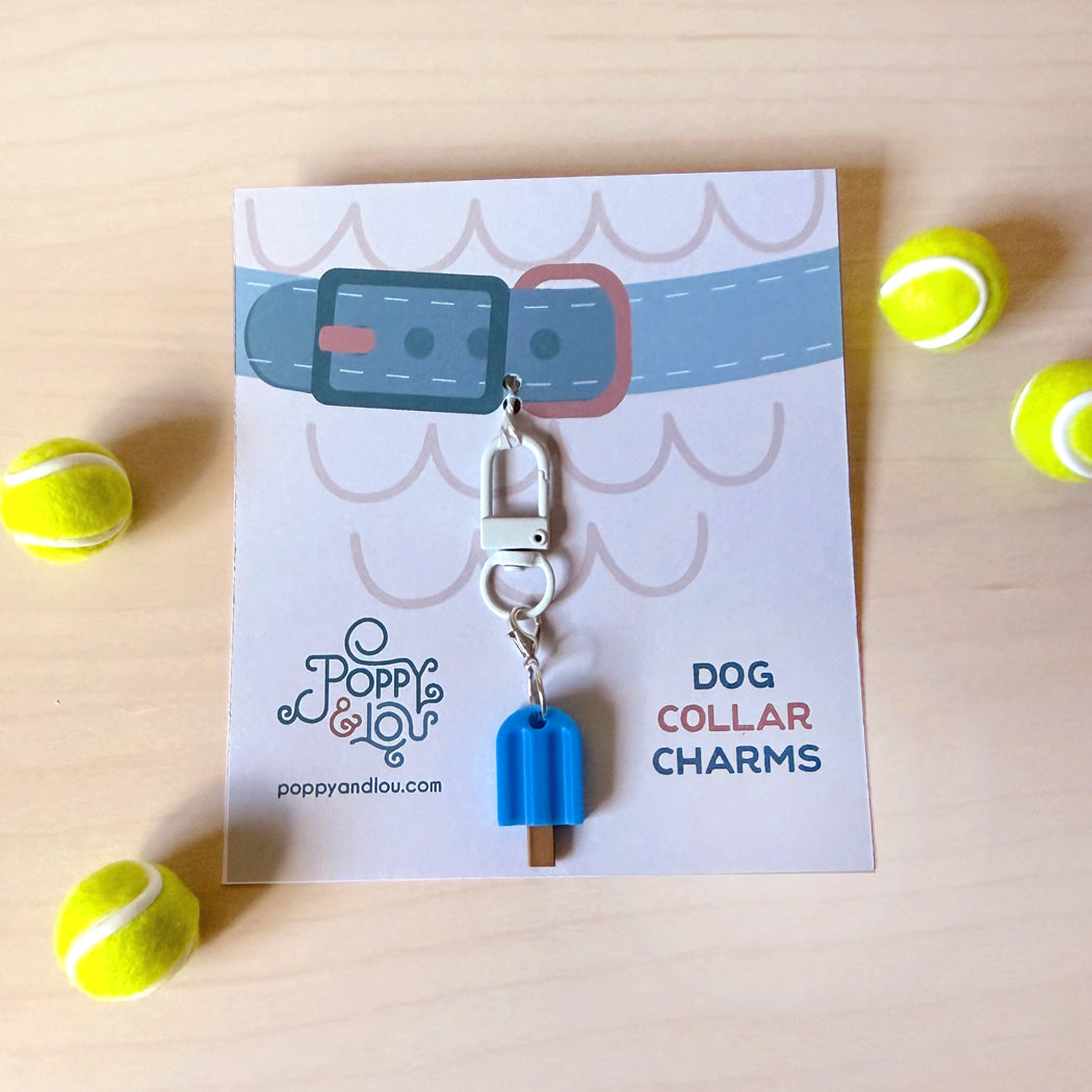 A blue popsicle-shaped dog collar charm with a silver clasp, displayed on a branded white card that reads 'DOG COLLAR CHARMS' and shows the Poppy & Lou brand. The card is photographed on a light wood surface with tennis balls around it.