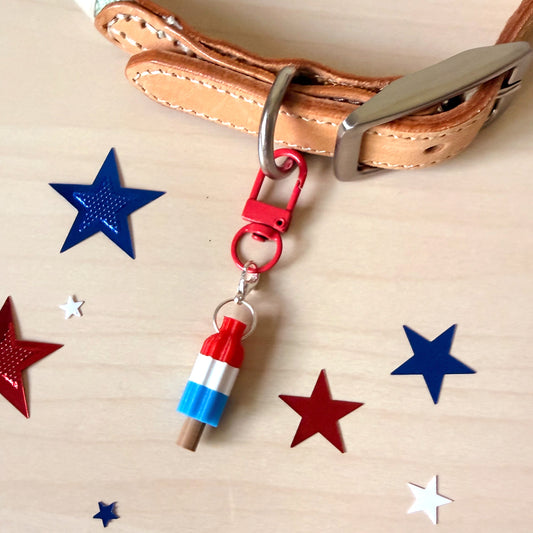 A patriotic-themed dog collar charm featuring a miniature red, white, and blue popsicle stick with a wooden dowel, attached to a red clasp and silver metal ring on a tan leather collar. The scene is styled on a light wood background with red, white, and blue star confetti.