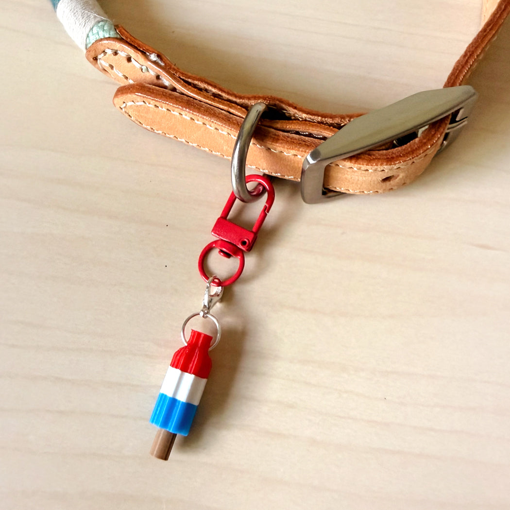 A close-up of a tan leather collar with a silver buckle and a red carabiner clip holding a small charm shaped like a striped popsicle in red, white, and blue, placed on a light wood surface.