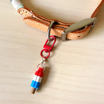 A close-up of a tan leather collar with a silver buckle and a red carabiner clip holding a small charm shaped like a striped popsicle in red, white, and blue, placed on a light wood surface.