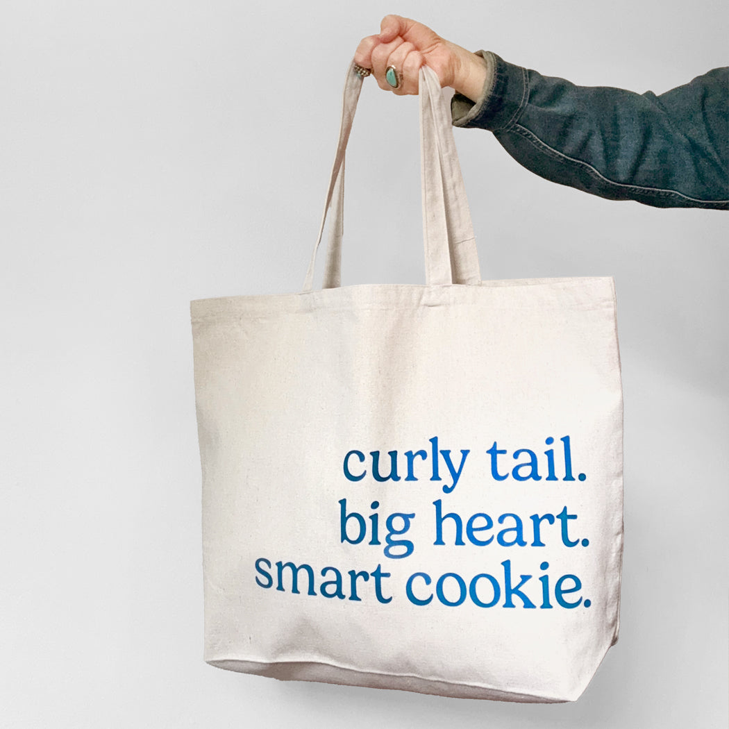 Tote bag with text 'curly tail. big heart. smart cookie.' held by a person against a light gray background.