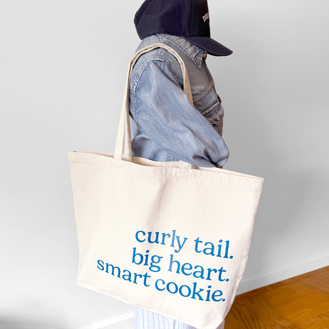 Person holding a tote bag with text 'curly tail. big heart. smart cookie.' against a plain background