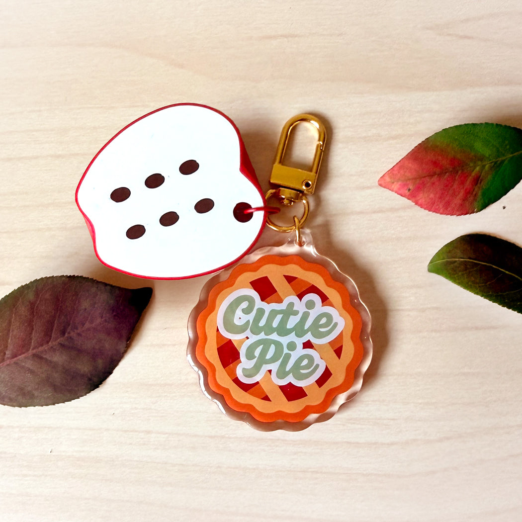A novelty keychain or bag charm set styled on a light wood surface with decorative leaves. One charm is shaped like a white slice of watermelon with red edges and black seeds, and the other is a clear acrylic pie charm with orange lattice crust, red filling, and the words 'Cutie Pie' in green script.