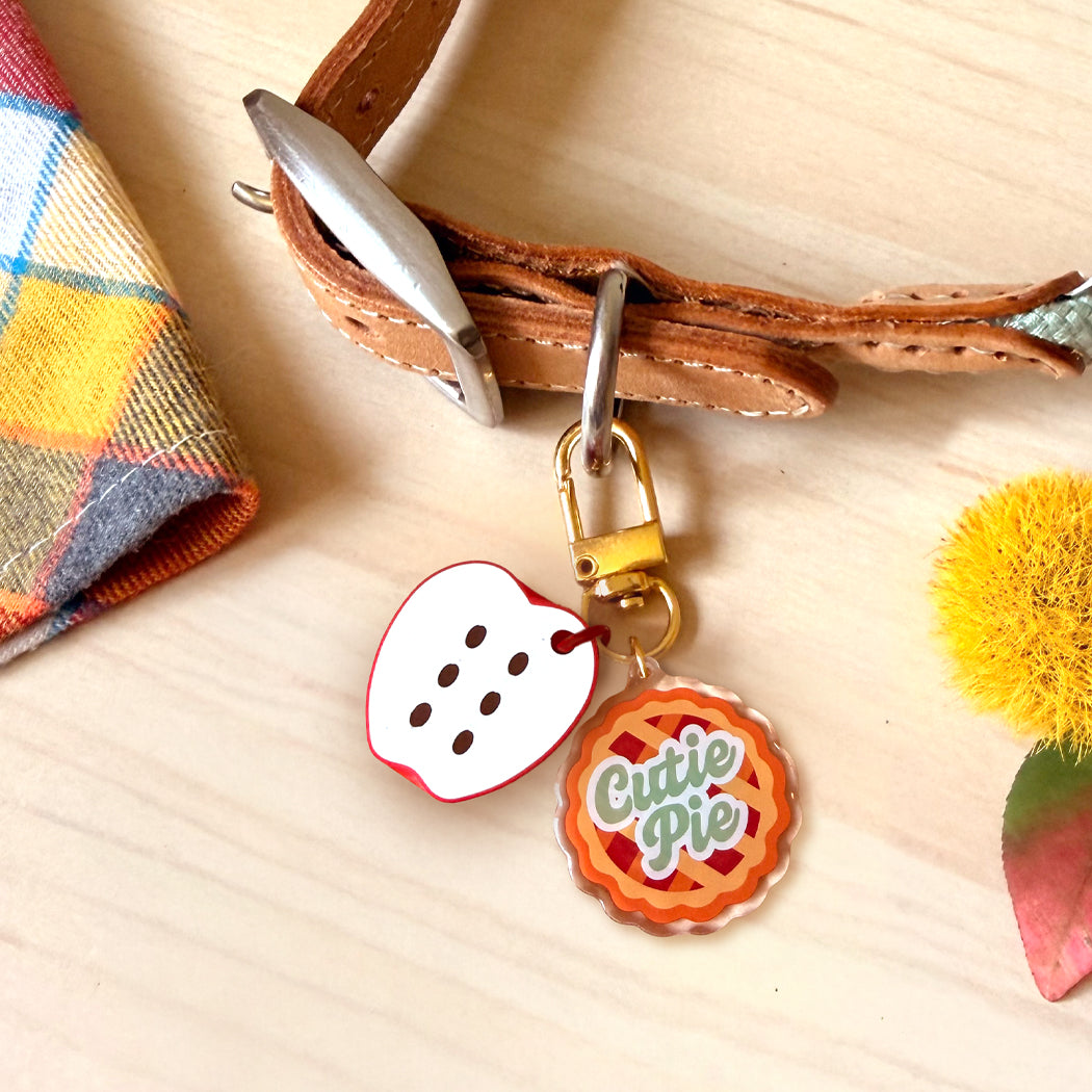 A close-up of a brown leather strap with metal hardware holding two decorative charms: a white heart-shaped charm with red edging and black dots, and a round orange-and-red pie charm with the words 'Cute Pie' in green script. The scene is styled on a light wood surface with a plaid fabric corner and a yellow pom-pom prop.