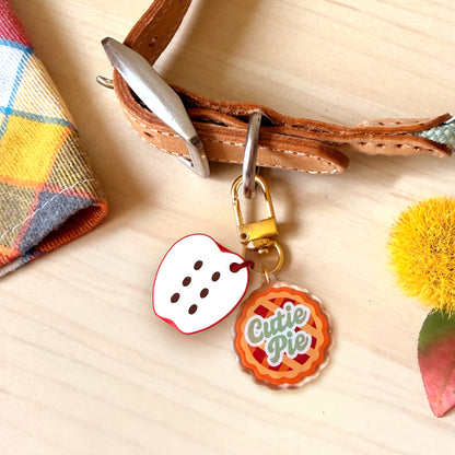 A close-up of a brown leather strap with metal hardware holding two decorative charms: a white heart-shaped charm with red edging and black dots, and a round orange-and-red pie charm with the words 'Cute Pie' in green script. The scene is styled on a light wood surface with a plaid fabric corner and a yellow pom-pom prop.