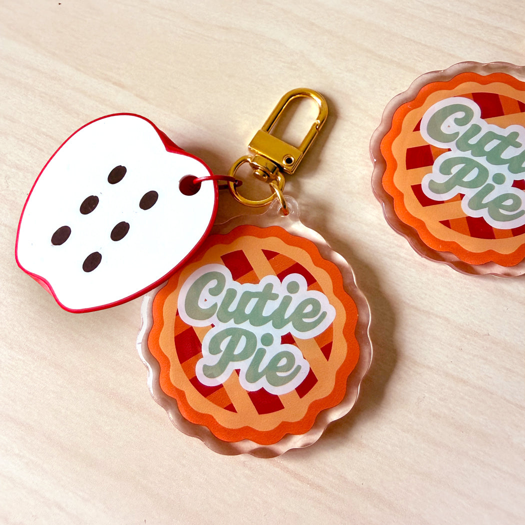 A set of acrylic keychains featuring a 'Cutie Pie' design and a white apple-shaped charm with black dots, attached to a gold key ring on a light wood background.