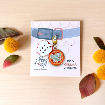 A product display card featuring two acrylic dog collar charms: a white die-cut charm with black dots and a red-and-green 'Cutie Pie' pie charm. The card is labeled 'Poppy andlou' and 'Dog Collar Charms', styled on a light wood background with decorative yellow pom-pom flowers and leaves.