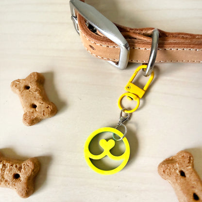 A tan leather dog collar with a silver buckle and a bright yellow keychain-style pet tag clasp, shown on a light wood surface with bone-shaped treats nearby.