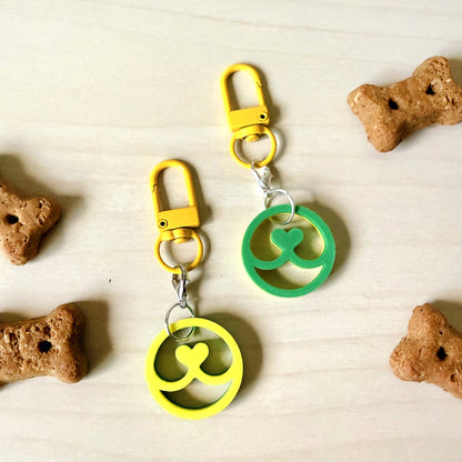 Two pet keychains with yellow and green circular charms shaped like a clover, attached to yellow clasps, displayed on a light wood surface with dog bone treats scattered around.