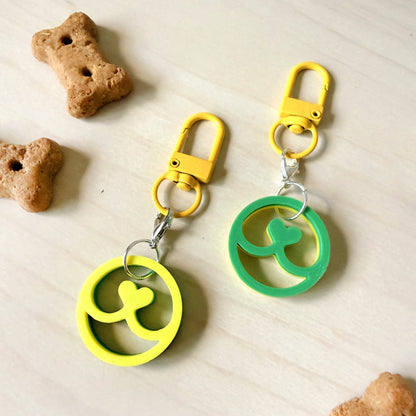 Two pet keychains with yellow and green peace sign charms, displayed on a light wood surface with dog treats as props.