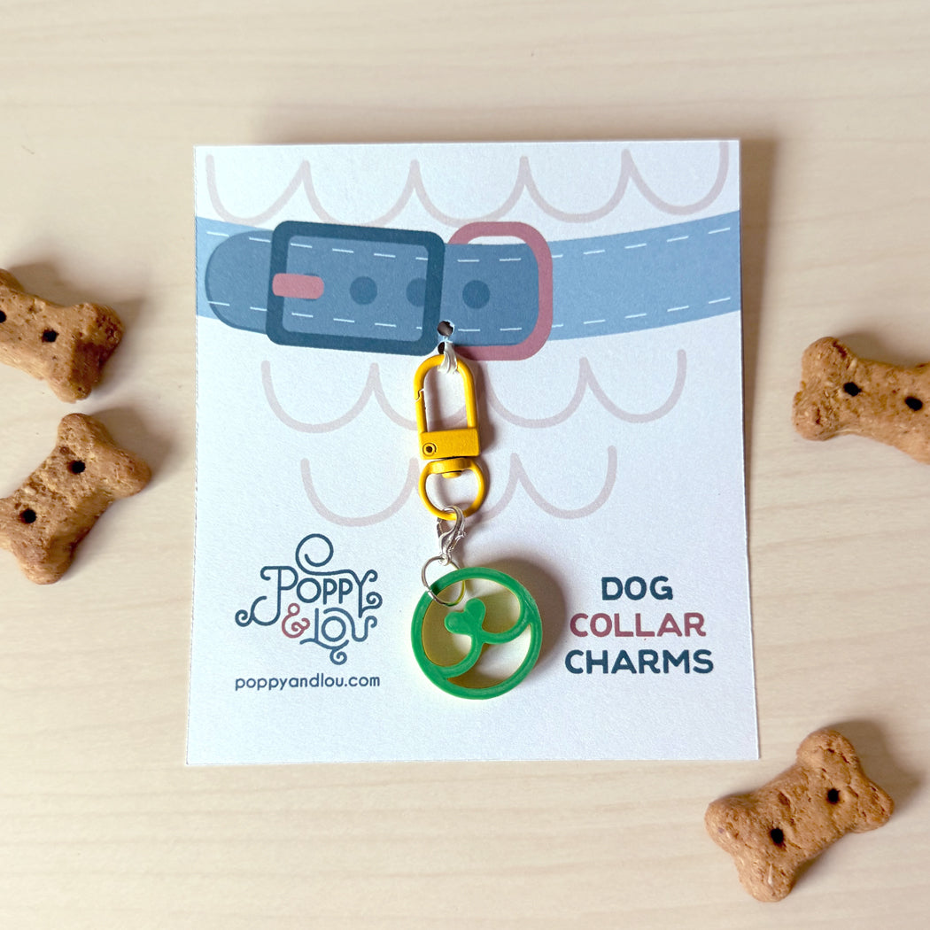 A green circular dog collar charm with a yellow carabiner clip, displayed on its printed backing card. The card features a blue and pink dog collar illustration and the brand name 'Poppy and Lou'.
