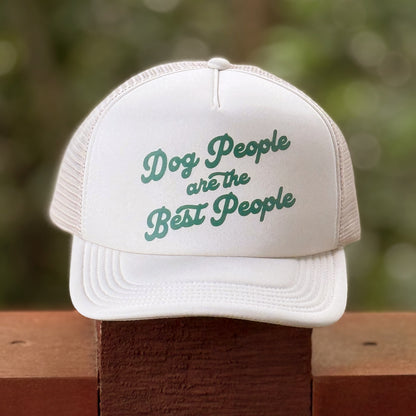 White cap with 'Dog People are the Best People' text on a wooden surface with a blurred green background