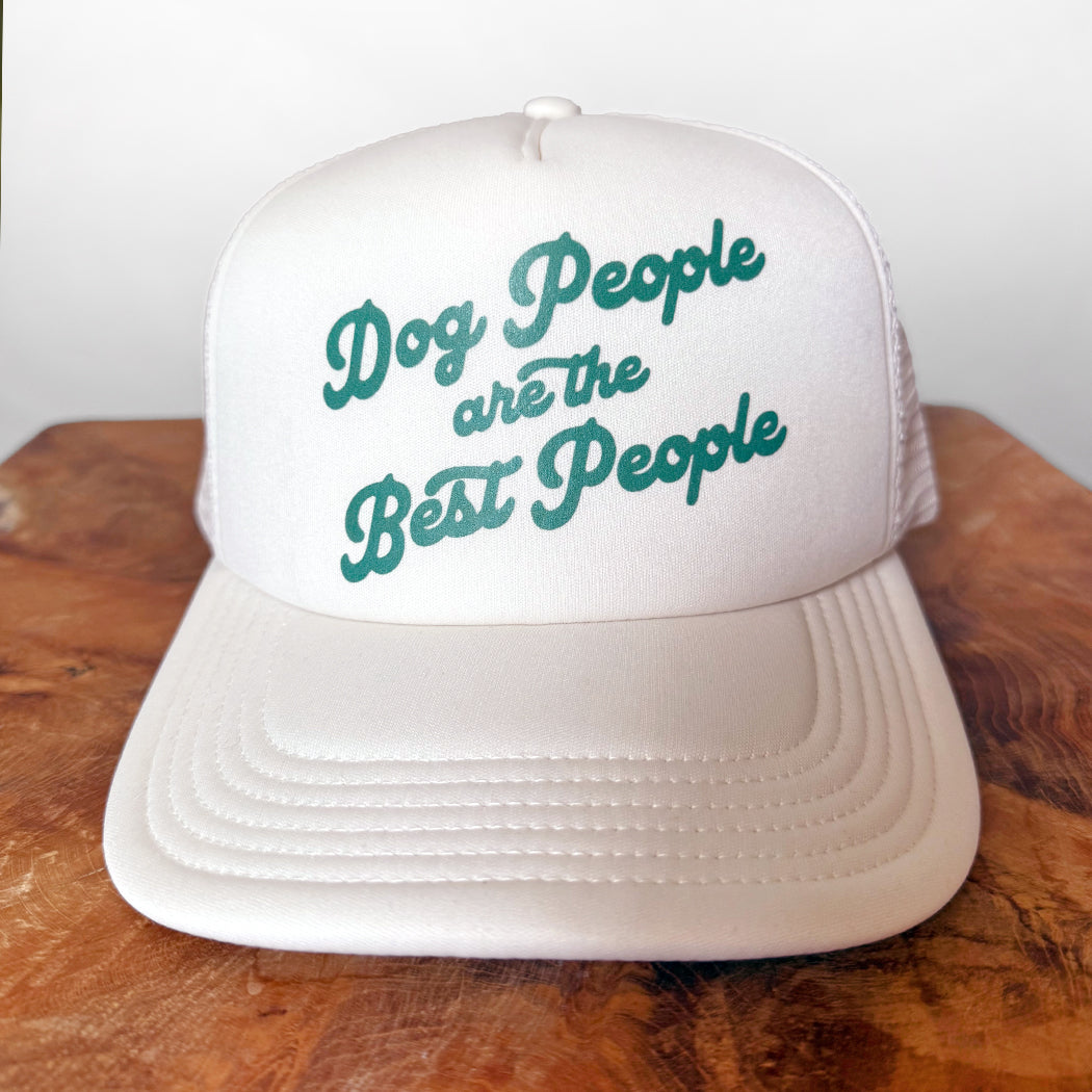 White cap with teal text 'Dog People are the Best People' on a wooden surface.