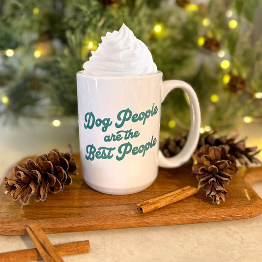 Mug with 'Dog People are the Best People' text, topped with whipped cream, on a wooden board with pinecones and cinnamon sticks, Christmas tree in the background.