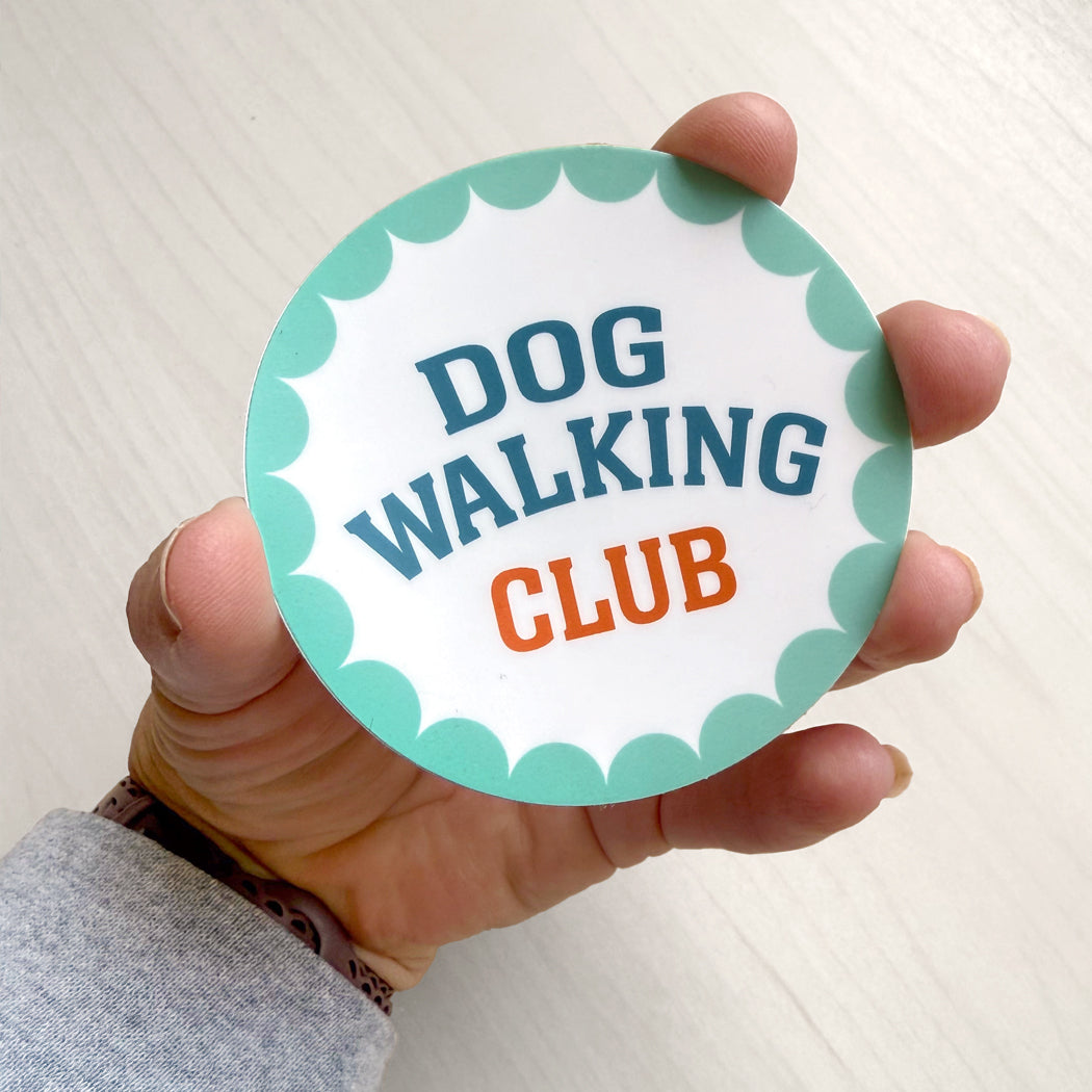 Hand holding a round vinyl Dog Walking Club sticker with teal scalloped border and bold text.