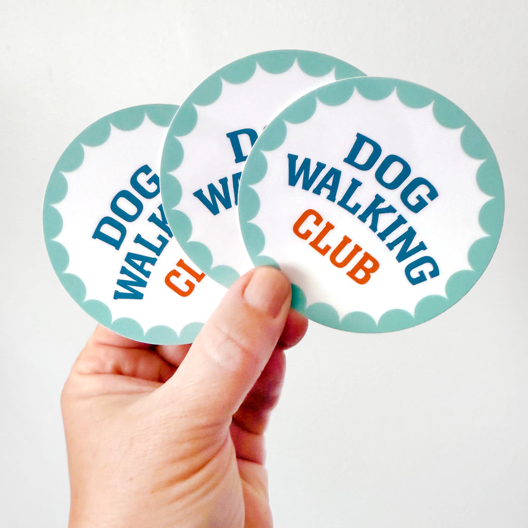 Hand holding a round vinyl Dog Walking Club sticker with teal scalloped border and bold text.