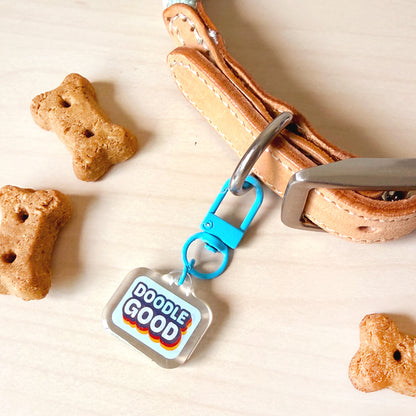 A close-up of a tan leather dog collar with a bright blue carabiner and a clear square tag that reads 'DOODLE GOOD' in colorful lettering. The scene is styled on a light wood surface with bone-shaped dog treats scattered around.
