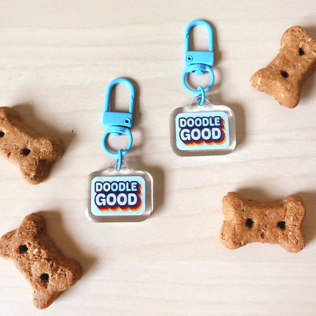 Two clear acrylic keychain tags with bright blue clips and a colorful 'DOODLE GOOD' logo, displayed on a light wood surface with decorative dog bone-shaped cookies around them.