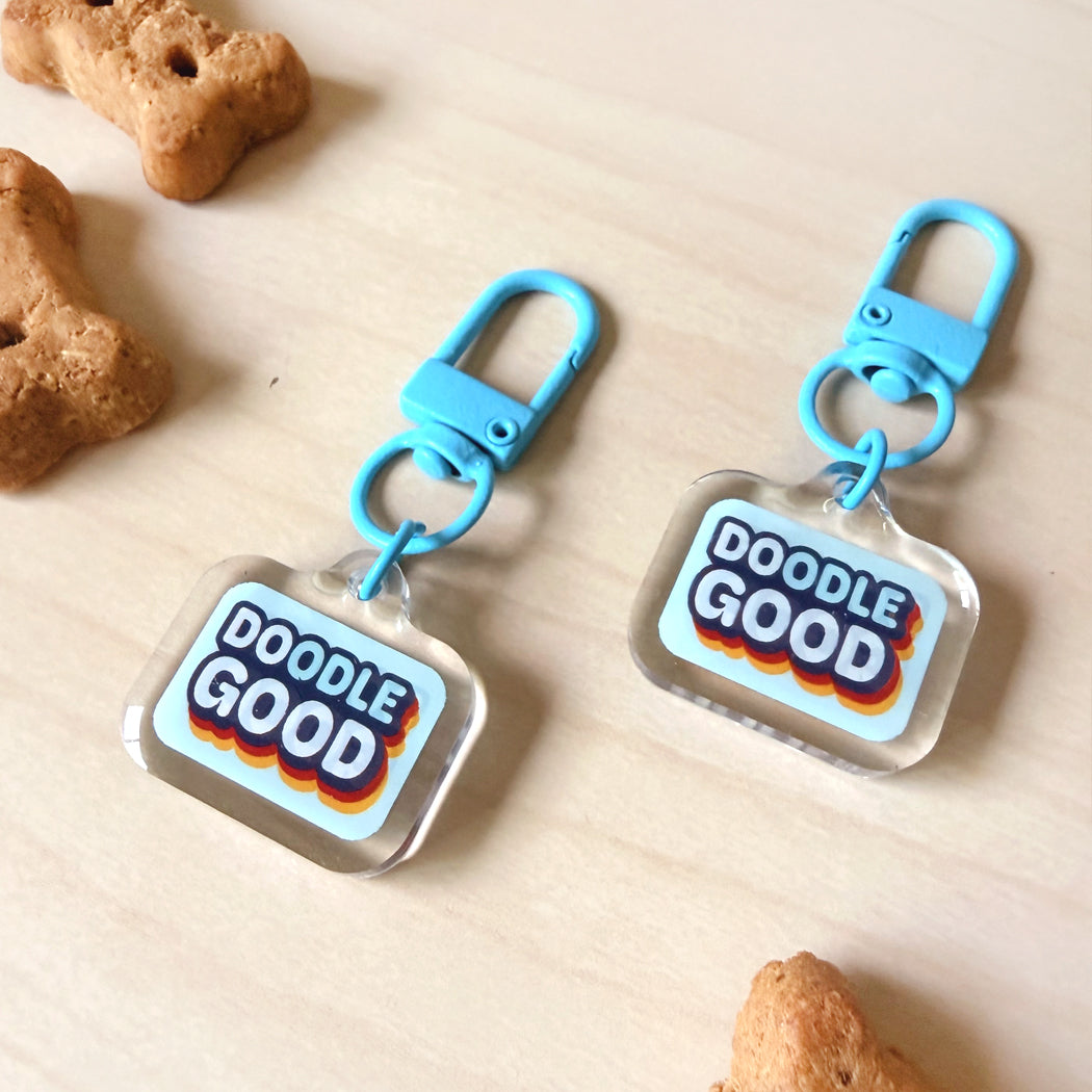 Two acrylic pet name tags with blue keychains, printed with 'DOODLE GOOD' in a retro style, displayed on a light wood surface with dog treats in the background.