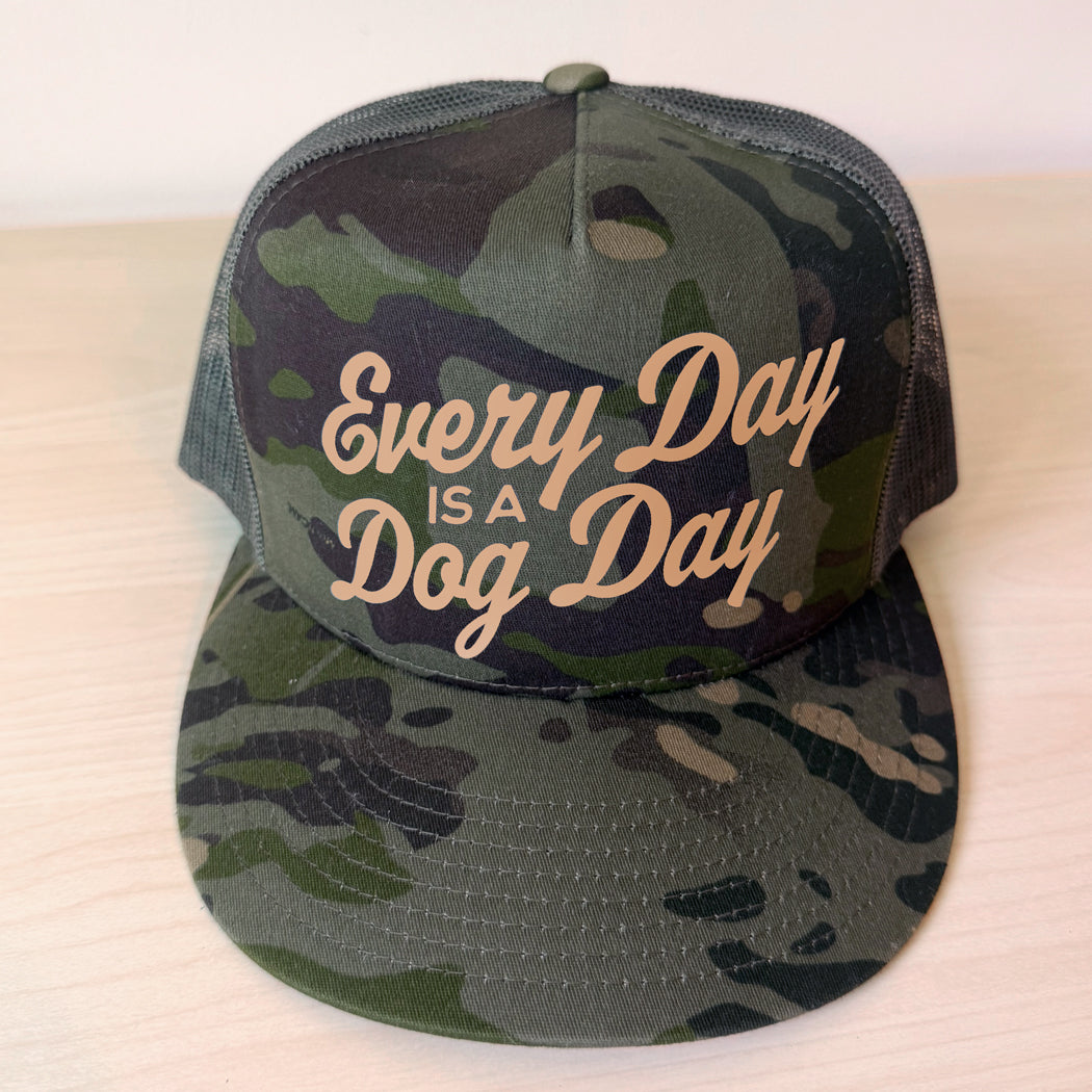 A camouflage trucker hat with mesh back panels and the phrase 'Every Day IS A Dog Day' in tan script on the front, displayed on a light wood surface.