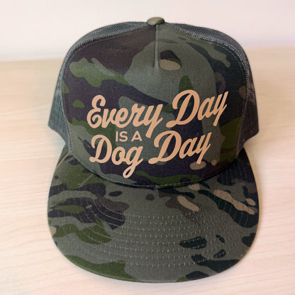 A camouflage trucker hat with mesh back panels and the phrase 'Every Day IS A Dog Day' in tan script on the front, displayed on a light wood surface.