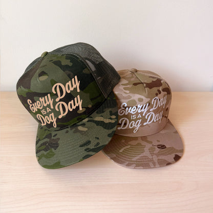 Two camouflage snapback caps on a light wood surface against a plain white wall. The front cap is green camo with white script text reading 'Every Day IS A Dog Day,' and the rear cap is tan camo with beige script text reading 'Every IS A Dog.