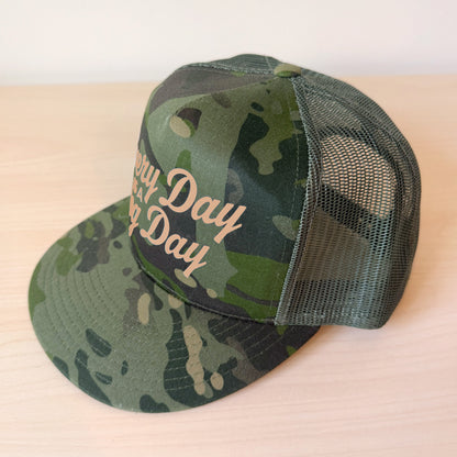 A side view of a dark green camouflage trucker cap with a curved brim and mesh back, featuring the text 'Every Day is a Dog Day' in tan script on the front panel, resting on a light wood surface.