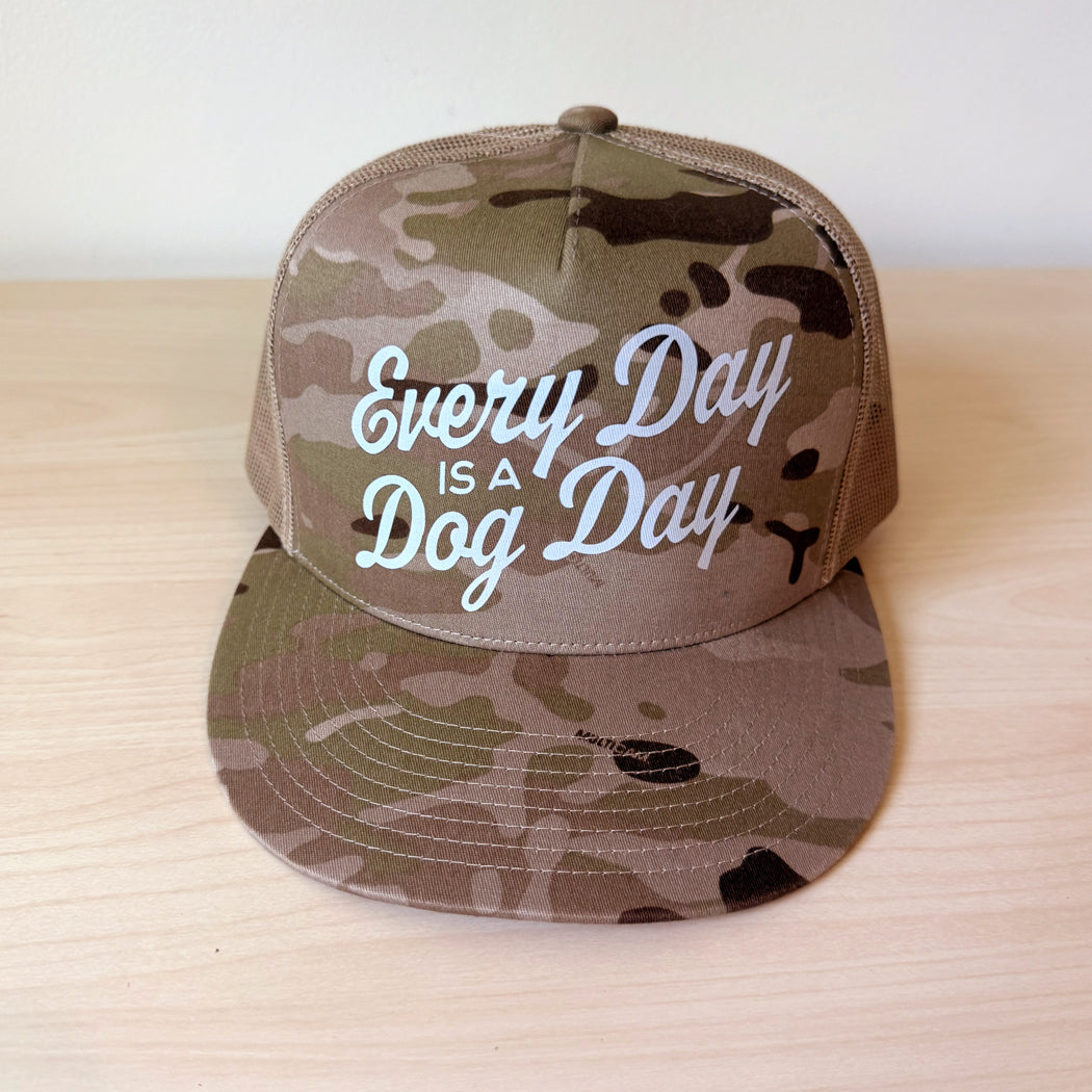 A camouflage tan trucker hat with mesh back panels and the phrase 'Every Day IS A Dog Day' in tan script on the front, displayed on a light wood surface.