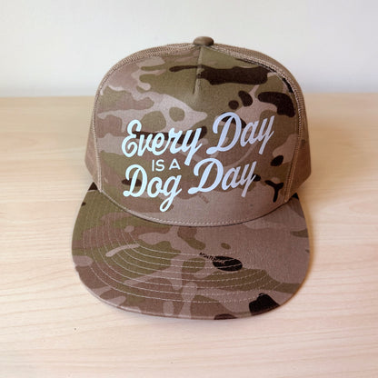 A camouflage tan trucker hat with mesh back panels and the phrase 'Every Day IS A Dog Day' in tan script on the front, displayed on a light wood surface.