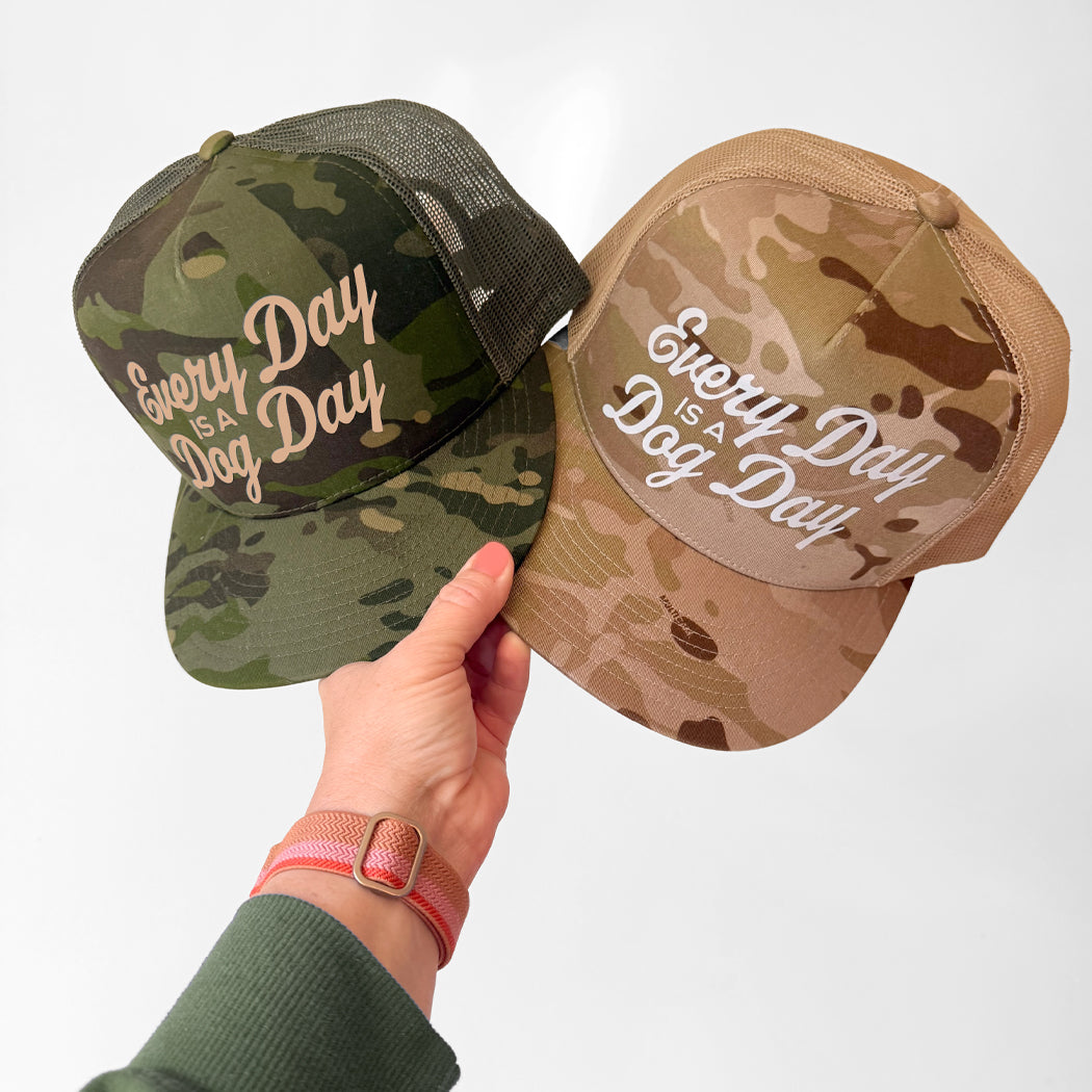 Two camouflage trucker hats held up by a hand against a plain light background. One hat is green woodland camo and the other is tan desert camo, both with white text reading 'Every Day is a Dog Day.