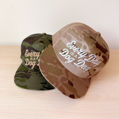 Two camouflage snapback caps on a light wood surface against a plain white wall. The front cap is tan and brown camo with white script text reading 'Every Day IS A Dog Day,' and the rear cap is dark green camo with beige script text reading 'Every IS A Dog.