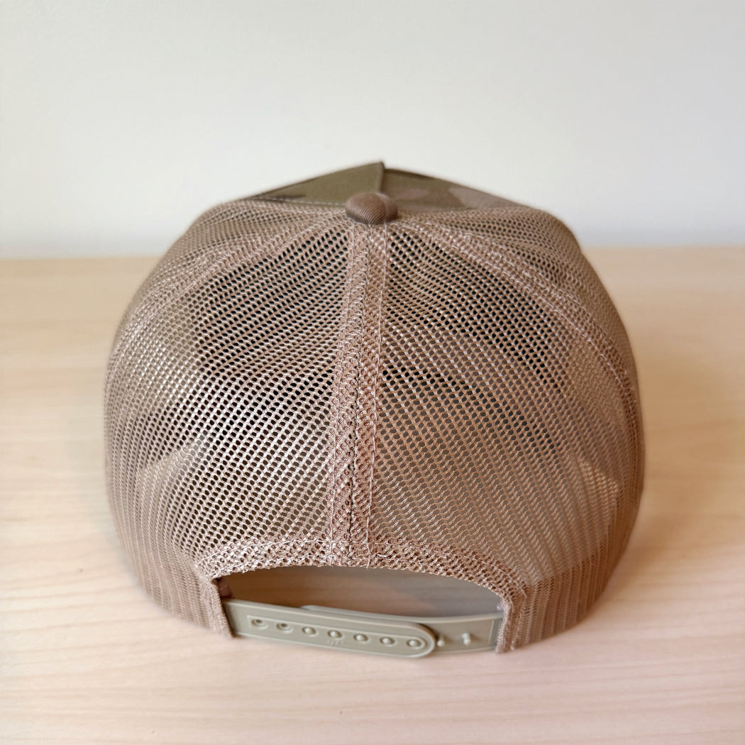 A camouflage trucker cap with a curved brim and mesh back, showing the back of teh hat resting on a light wood surface.
