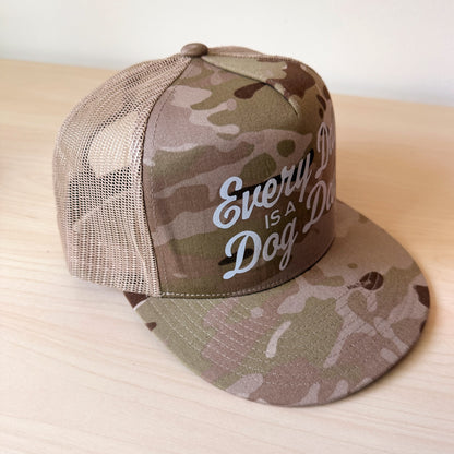 A tan camouflage trucker cap with a curved brim and mesh back, featuring the text 'Every Day is a Dog Day' in white script on the front panel, resting on a light wood surface.