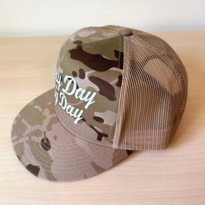 A camouflage trucker cap with a curved brim and mesh back, featuring the text 'Every Day is a Dog Day' in white script on the front panel, resting on a light wood surface.